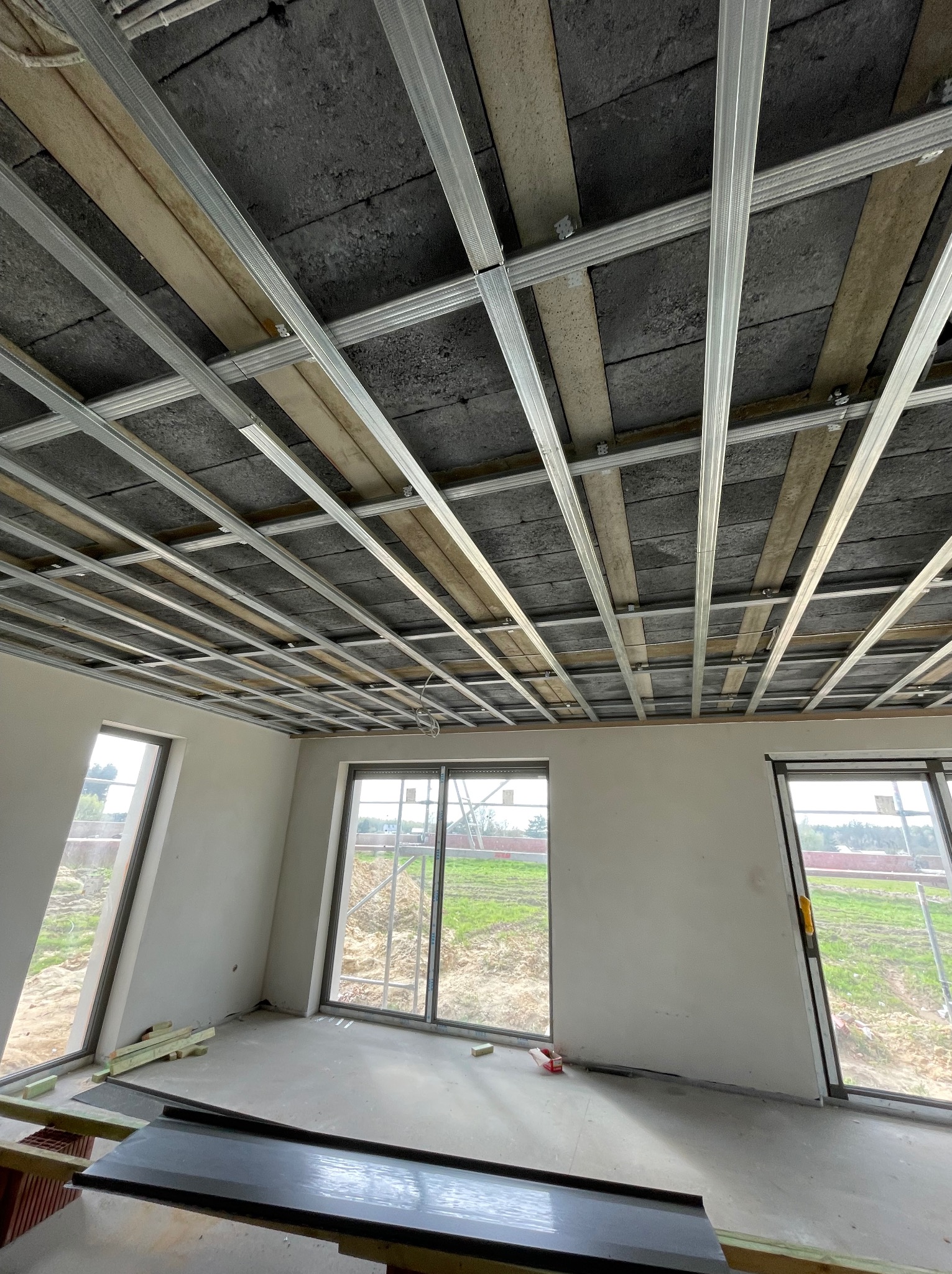 Metalowa konstrukcja sufitu podwieszanego w jasnym wnętrzu z dużymi oknami. Widoczne drewniane elementy konstrukcyjne i surowy betonowy strop.