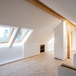 Marcin-Bau - Poddasze w trakcie prac wykończeniowych: surowe drewno, białe ściany, okna dachowe z widokiem na drzewa, podłoga z płyt OSB, elementy zabudowy.