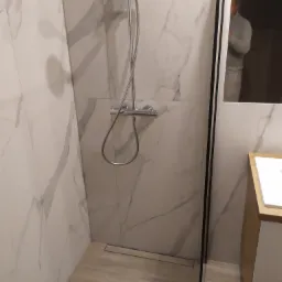 Nowoczesna kabina prysznicowa z odpływem liniowym, wykończona białymi płytkami imitującymi marmur, z widoczną armaturą i fragmentem szafki umywalkowej.