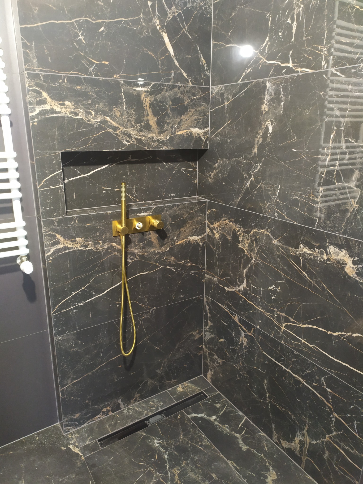 Nowoczesna kabina prysznicowa z czarnymi płytkami imitującymi marmur, złotą armaturą i odpływem liniowym w podłodze.