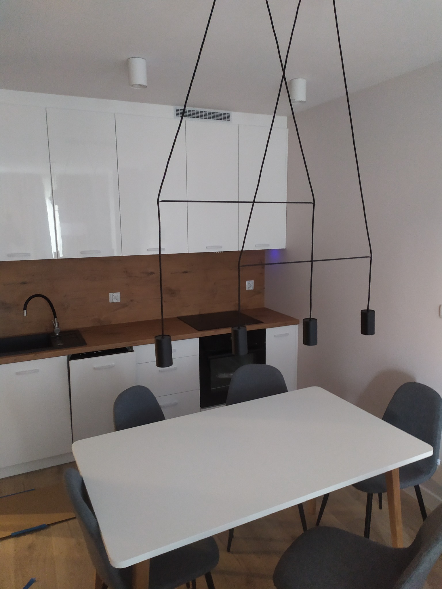 Nowoczesna kuchnia z białymi szafkami, drewnianym blatem, czarnym zlewem i minimalistyczną lampą wiszącą nad białym stołem z szarymi krzesłami.