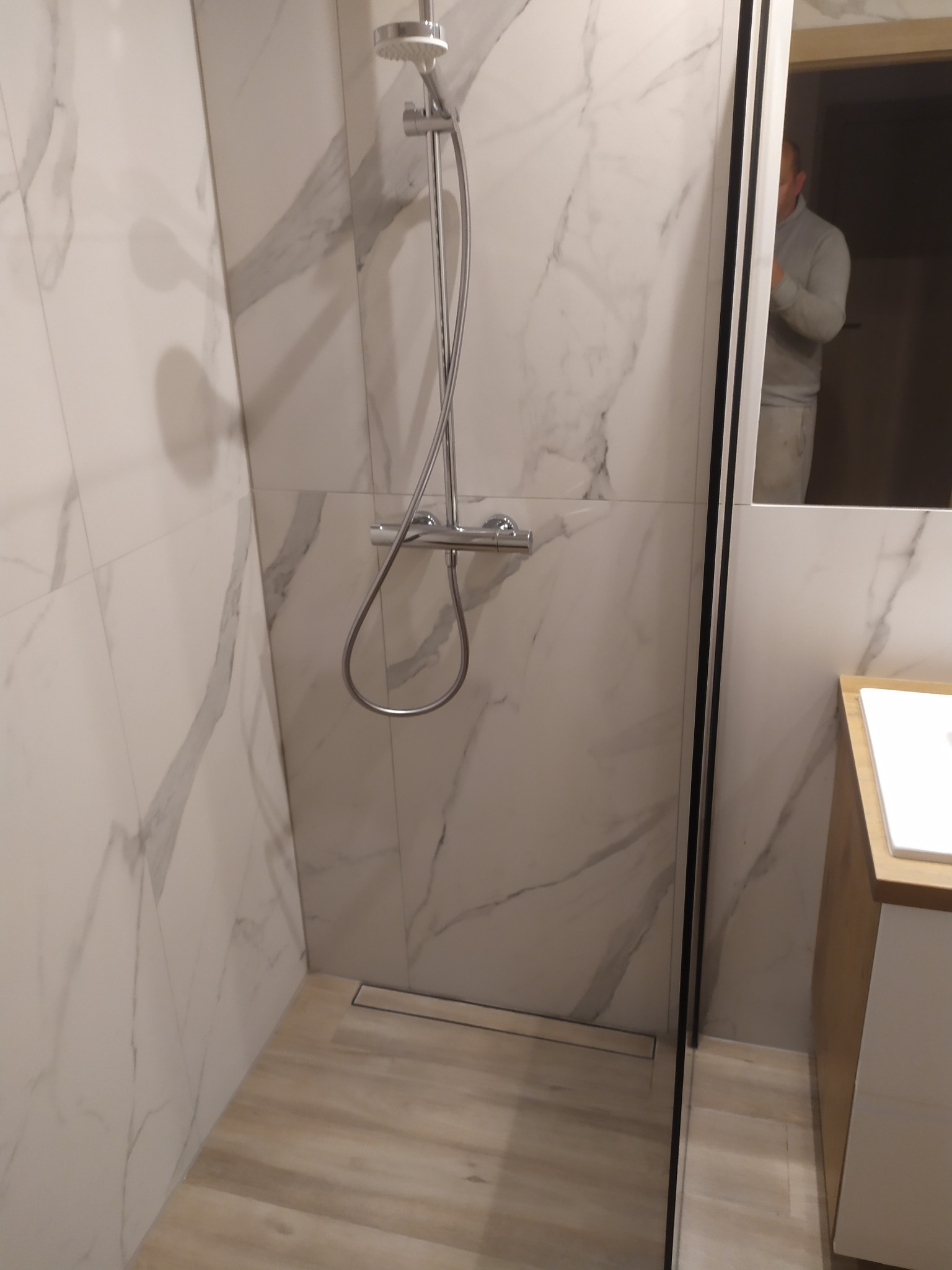 Nowoczesna kabina prysznicowa z odpływem liniowym, wykończona białymi płytkami imitującymi marmur, z widoczną armaturą i fragmentem szafki umywalkowej.