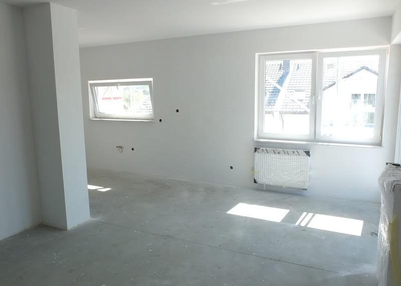 Surowe wnętrze pokoju z białymi ścianami, betonową podłogą, dwoma oknami i grzejnikiem zabezpieczonym folią, przygotowane do dalszych prac wykończeniowych.