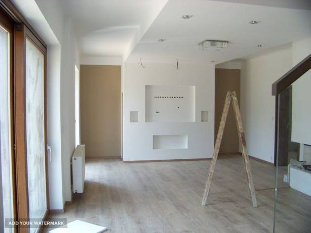 Jasne, przestronne wnętrze w trakcie prac wykończeniowych, z drewnianą podłogą, białą ścianą z wnękami, grzejnikiem pod oknem i drewnianą drabiną pośrodku pomieszczenia.