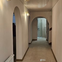 ProBau K&W - Korytarz z beżowymi płytkami podłogowymi, białymi ścianami z łukowymi przejściami i kryształowymi kinkietami, widok na łazienkę w głębi.