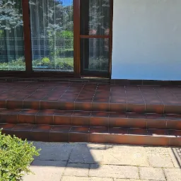 Kafelkowane schody wejściowe do domu z brązowymi płytkami, widok z dołu. Na pierwszym planie krzew, w tle okno z zasłonami i fragment elewacji.