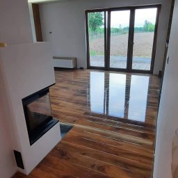 ProBau K&W - Salon z błyszczącą, drewnopodobną podłogą, kominkiem i dużym oknem tarasowym. Widok na zewnątrz. Nowoczesne wykończenie wnętrza, minimalistyczny design.