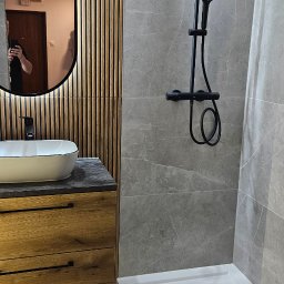 ProBau K&W - Nowoczesna łazienka z drewnianymi lamelami, szarą płytką imitującą marmur i czarną armaturą. Umywalka nablatowa i kabina prysznicowa z brodzikiem.