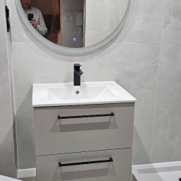 ProBau K&W - Nowoczesna łazienka w szarościach: umywalka z szafką, okrągłe lustro z podświetleniem i kabina prysznicowa. W lustrze odbija się osoba robiąca zdjęcie.