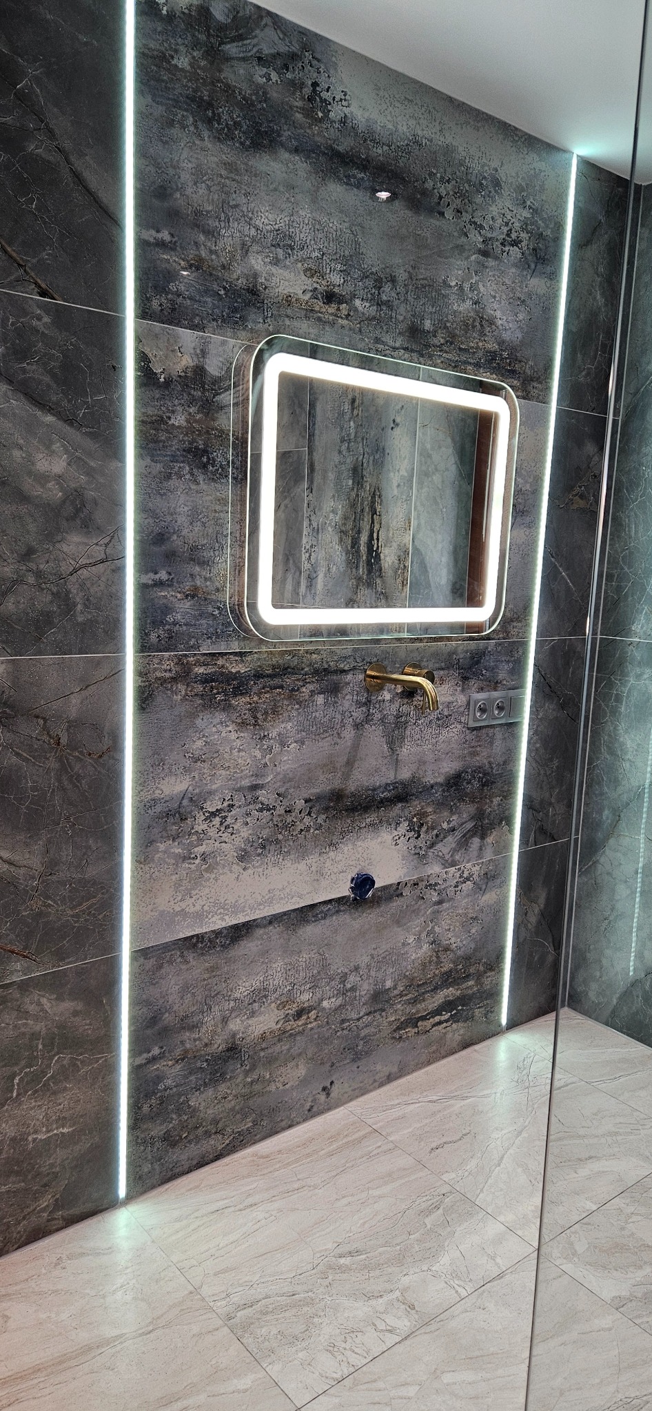 Wykończona łazienka z szarymi płytkami imitującymi kamień, podświetlanym lustrem i złotą armaturą, widoczny fragment szklanej kabiny prysznicowej.