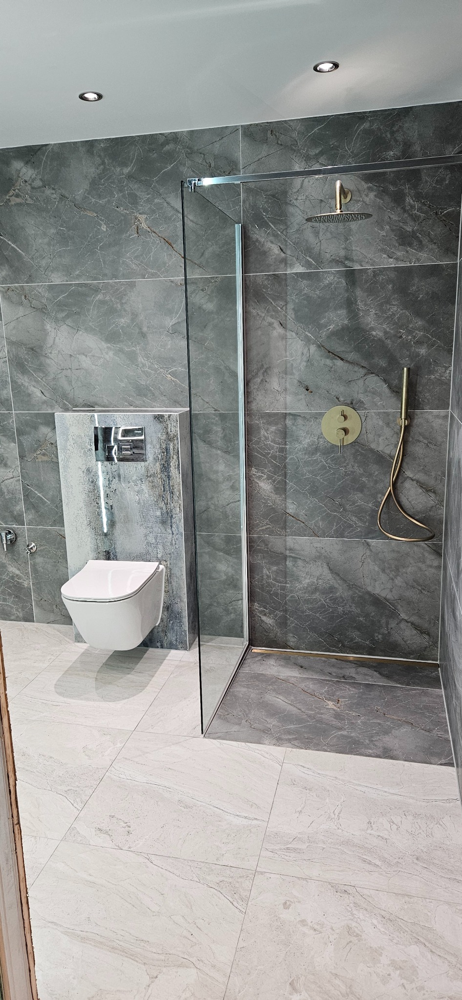 Nowoczesna łazienka z szarymi płytkami imitującymi marmur, wiszącą toaletą i szklaną kabiną prysznicową z złotą armaturą.