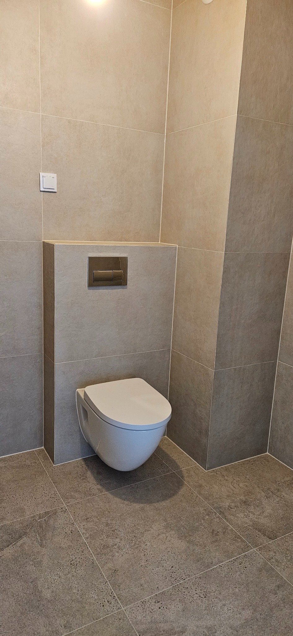 Podwieszana toaleta w jasnej łazience wykończonej płytkami w odcieniach beżu i szarości. Widoczny przycisk spłukiwania i fragment podłogi z płytek.