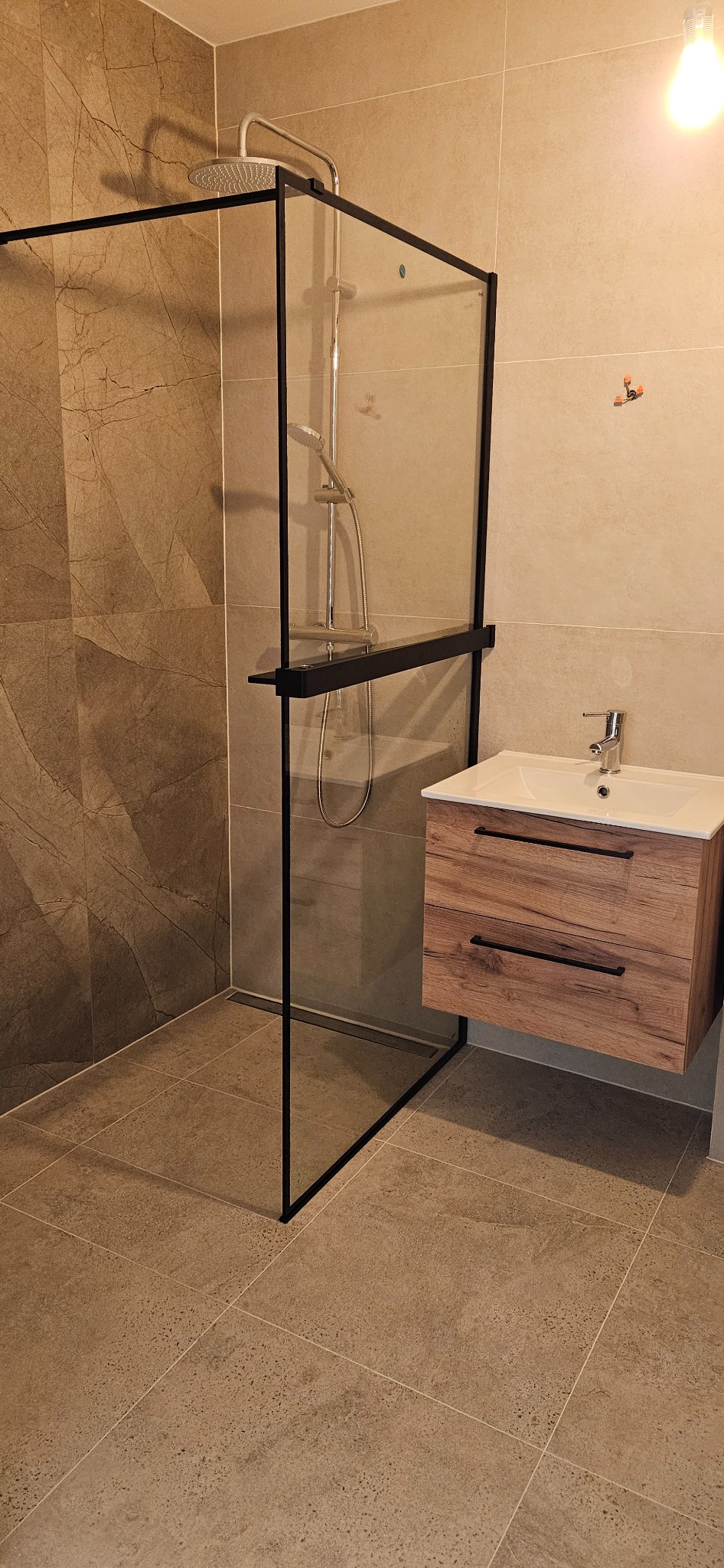 Nowoczesna łazienka z kabiną typu walk-in z czarnymi profilami, drewnianą szafką pod umywalką i płytkami imitującymi kamień w beżowych odcieniach.