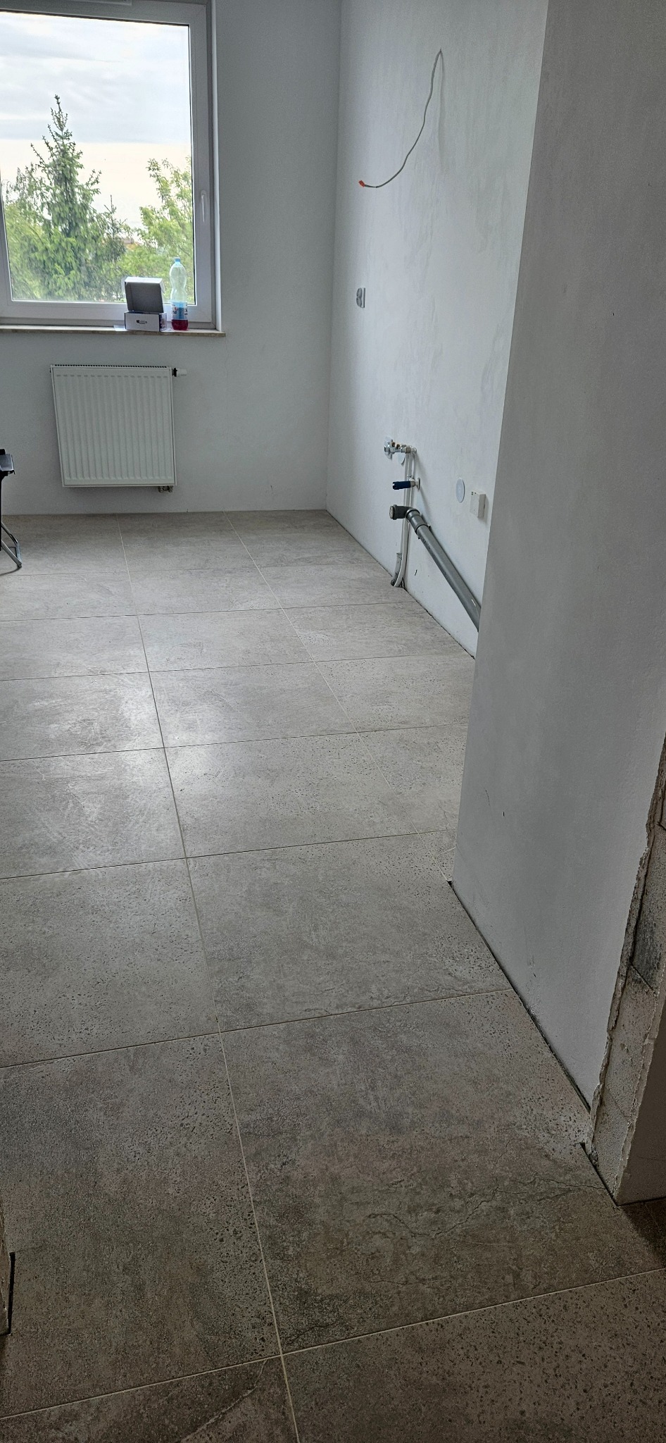 Beżowe płytki podłogowe, świeżo położone w pustym pomieszczeniu z grzejnikiem i instalacją na ścianie. Widok na okno z drzewami w tle, neutralne oświetlenie.