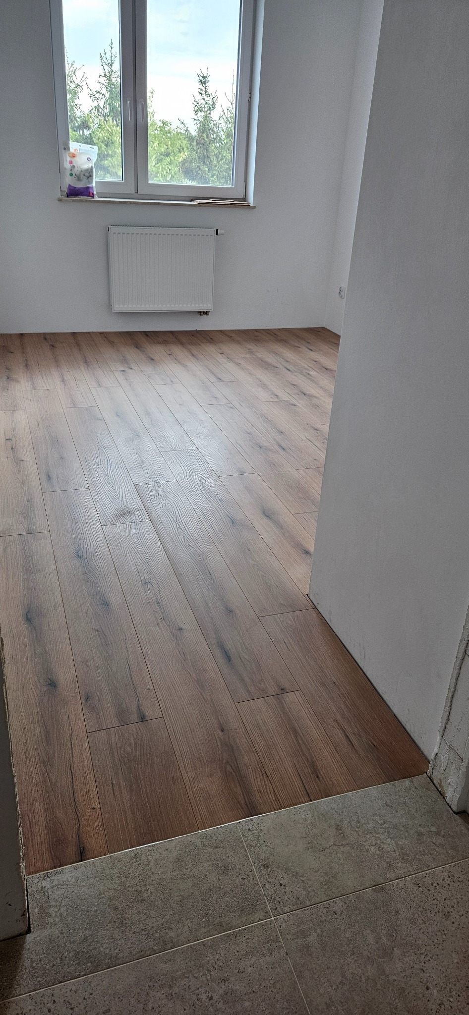 Ułożony parkiet laminowany w nowym, jasnym pokoju z białymi ścianami, widok z progu na okno. Podłoga imitująca drewno, w tle grzejnik i okno z widokiem na zieleń.