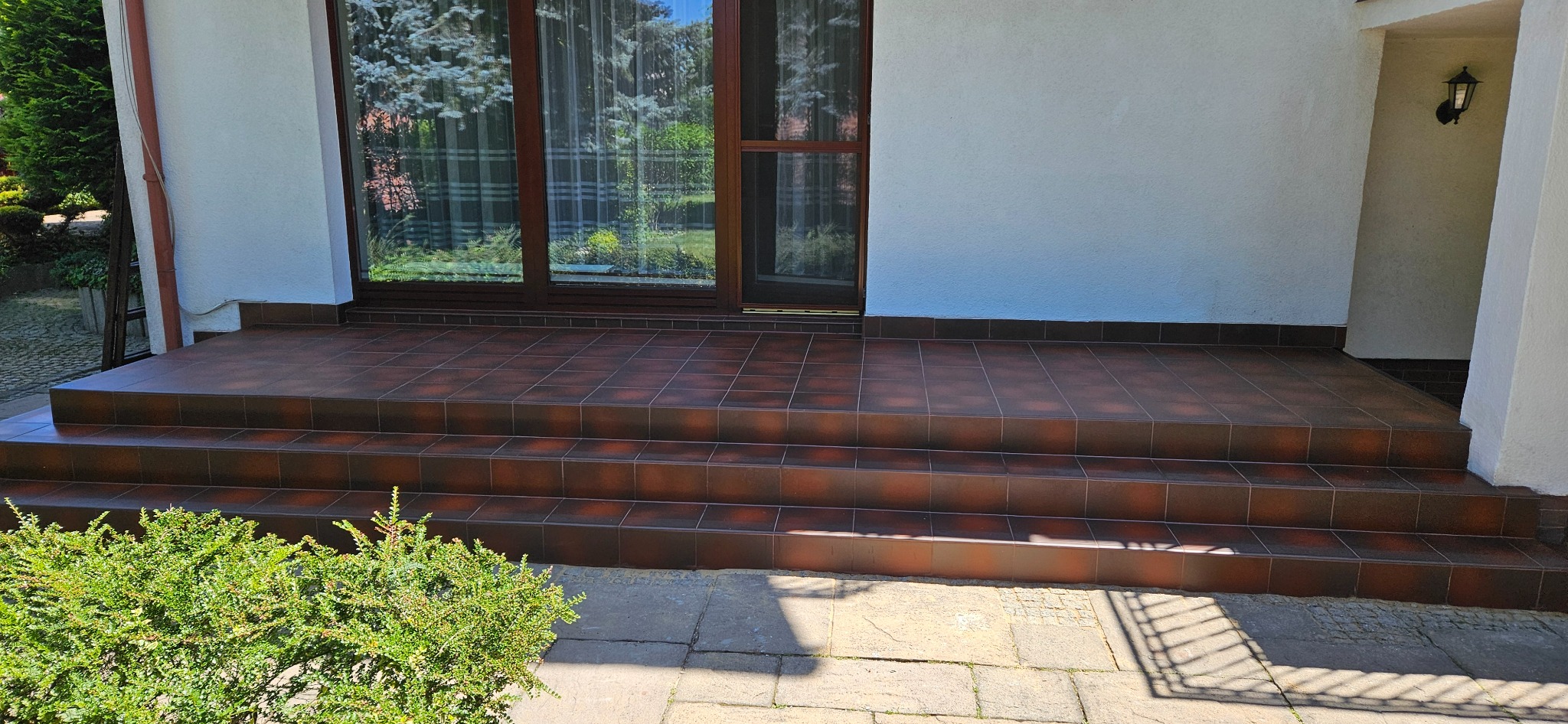 Kafelkowane schody wejściowe do domu z brązowymi płytkami, widok z dołu. Na pierwszym planie krzew, w tle okno z zasłonami i fragment elewacji.