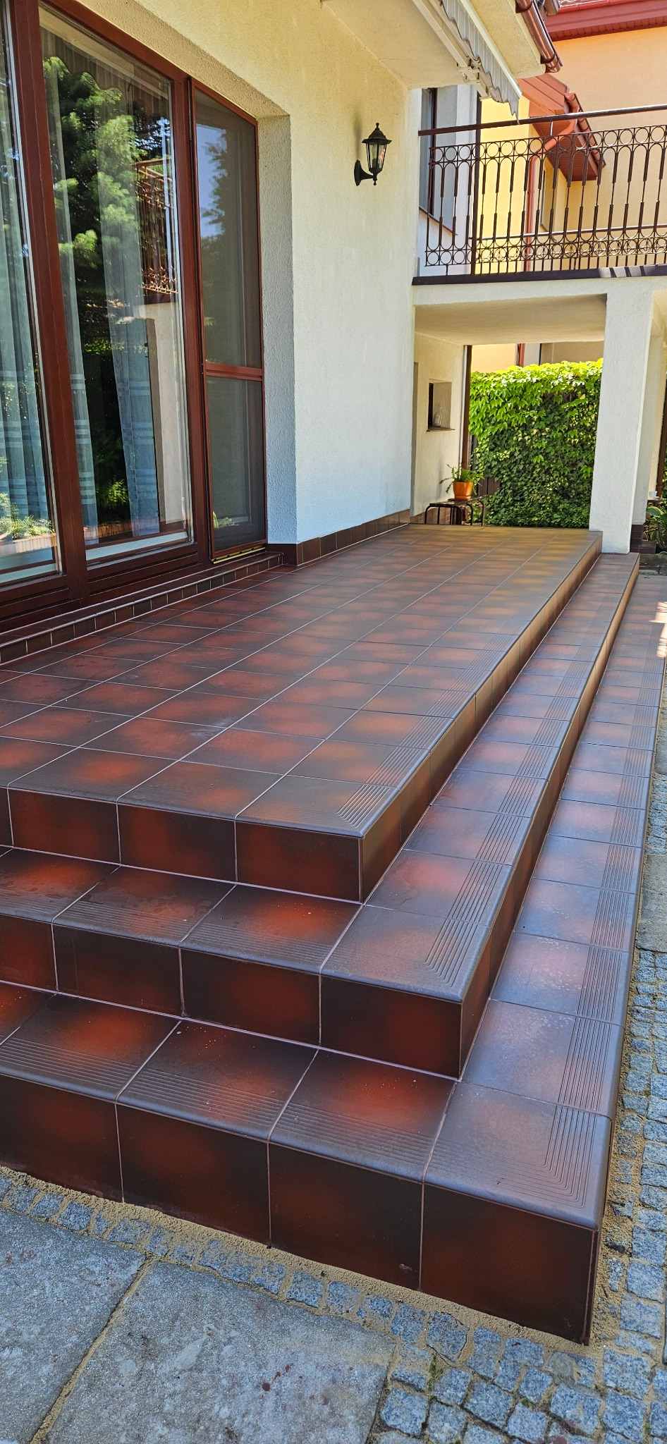 Brązowe płytki na schodach zewnętrznych prowadzących do domu. Widoczne detale wykończenia i fuga. Schody z płytek na zewnątrz budynku.