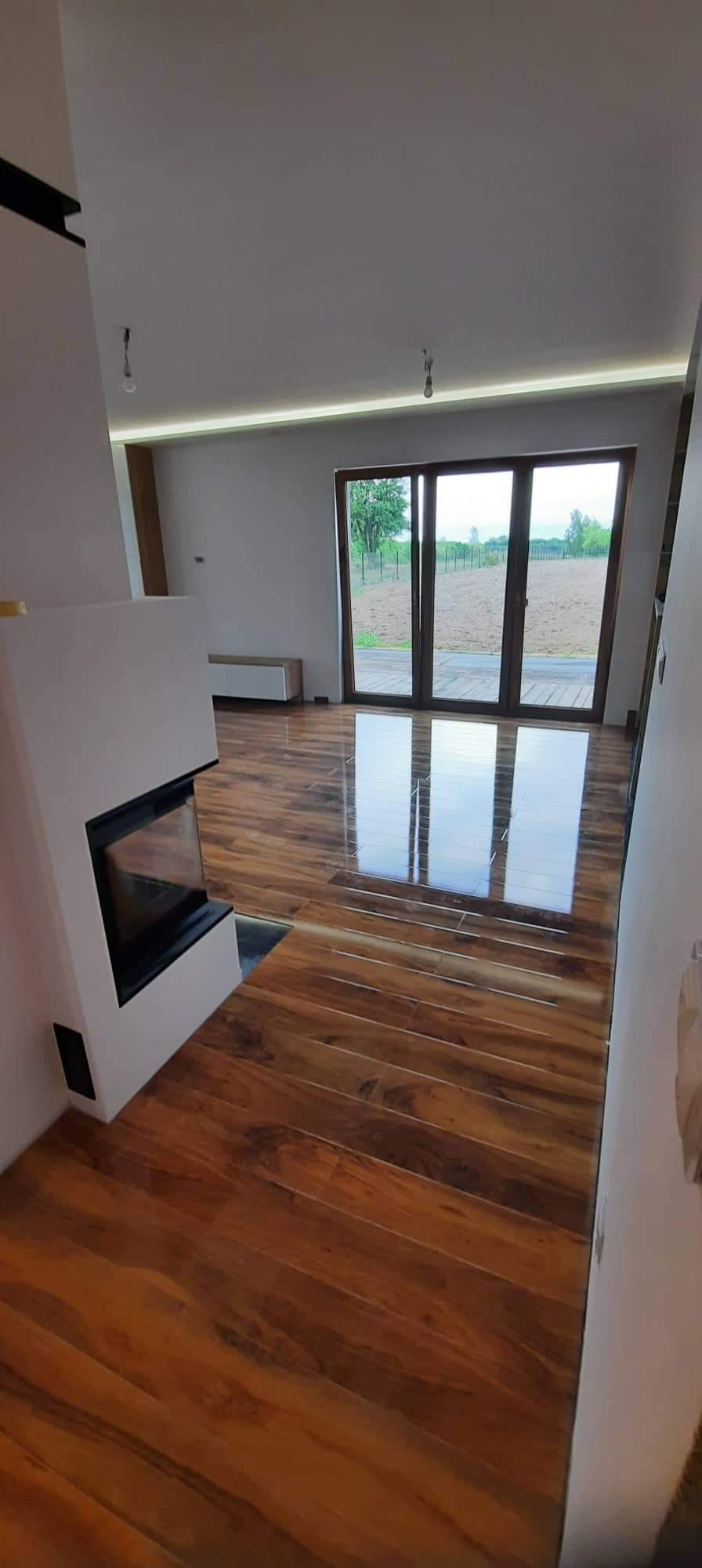 Salon z błyszczącą, drewnopodobną podłogą, kominkiem i dużym oknem tarasowym. Widok na zewnątrz. Nowoczesne wykończenie wnętrza, minimalistyczny design.