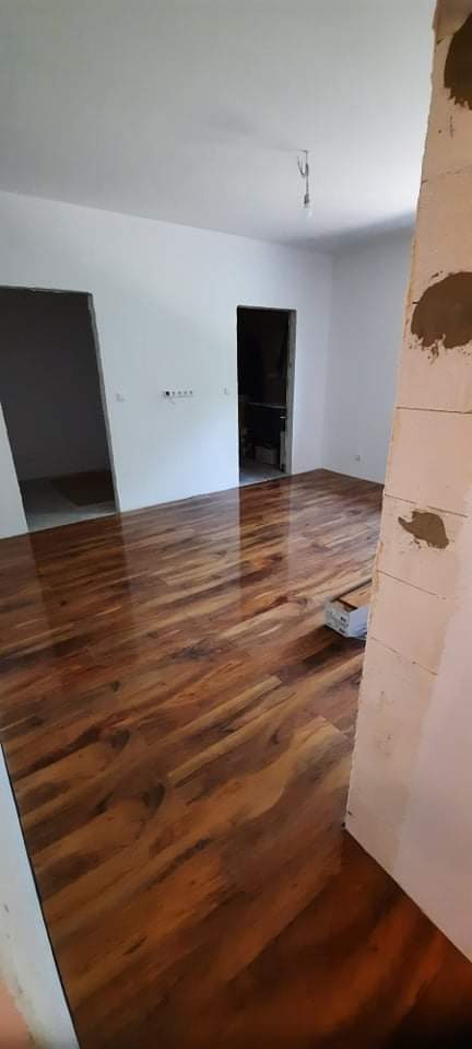 Połyskująca, nowa drewniana podłoga w jasnym pomieszczeniu z dwoma otworami drzwiowymi i niedokończoną ścianą. Widoczne paczki paneli podłogowych.