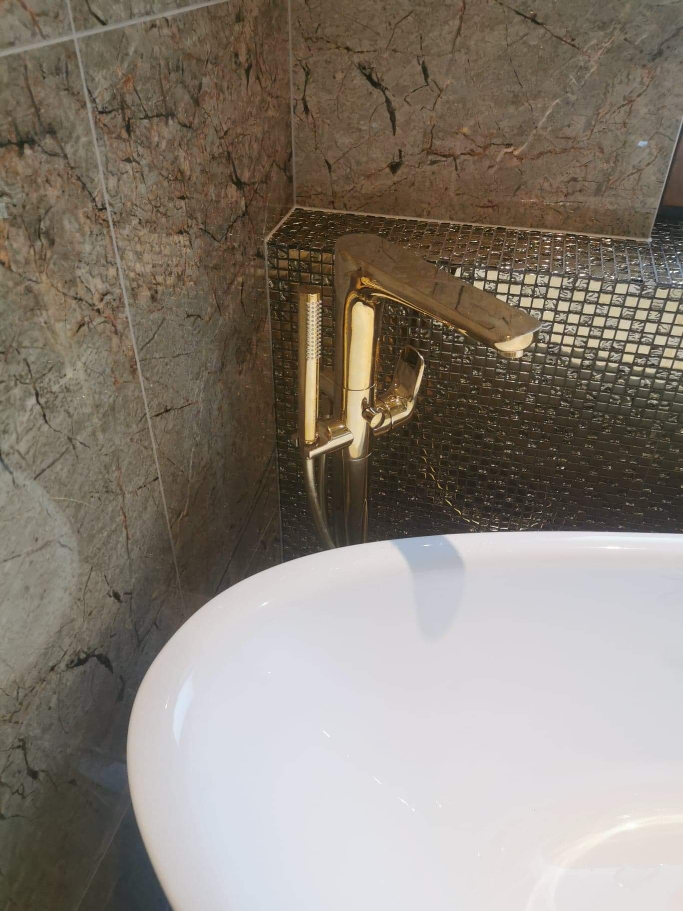 Bateria wannowa w kolorze złotym na tle mozaiki i płytek imitujących marmur. Element wykończenia łazienki, nowoczesny design.