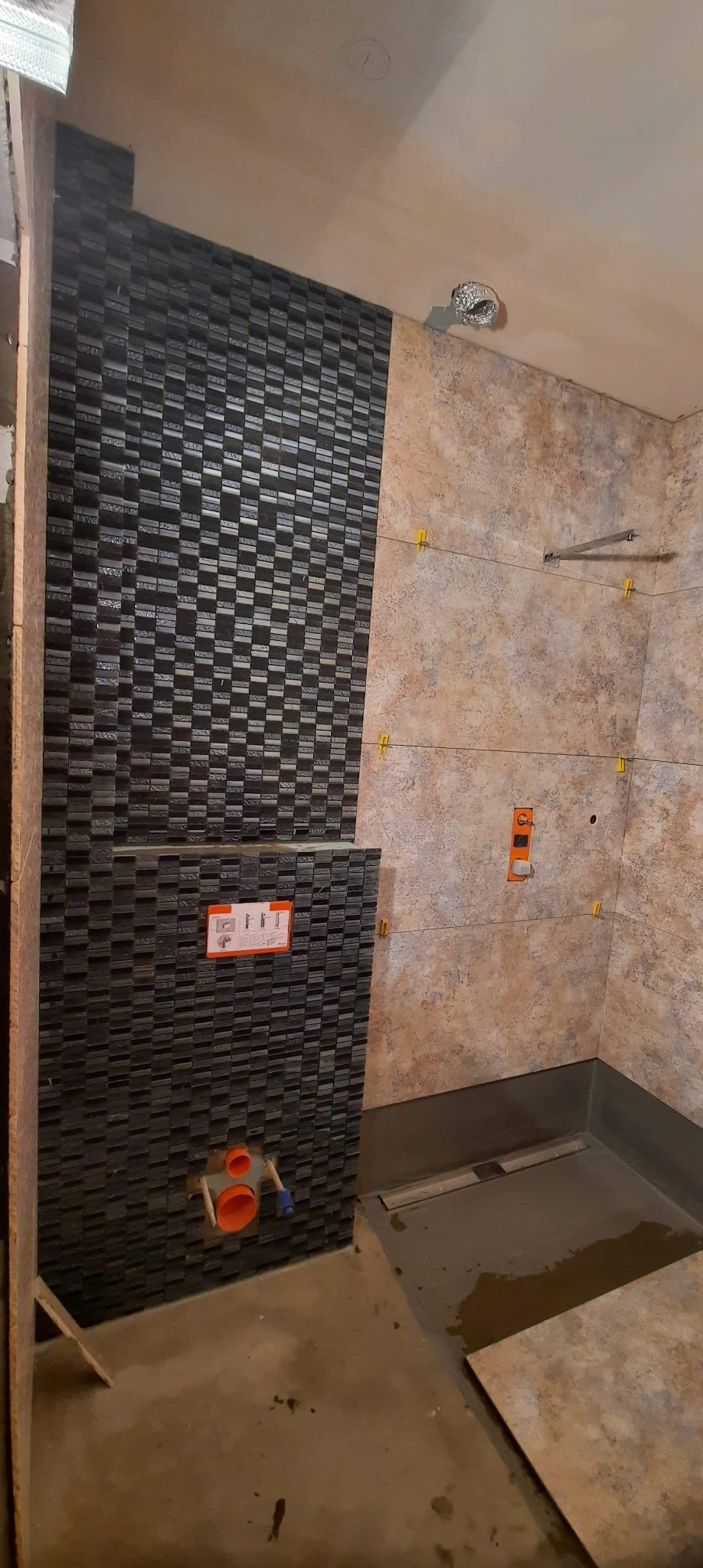 Łazienka w trakcie wykańczania: częściowo ułożone płytki mozaikowe w odcieniach szarości, beżowe płytki na ścianie, widoczne elementy instalacji i poziomica.