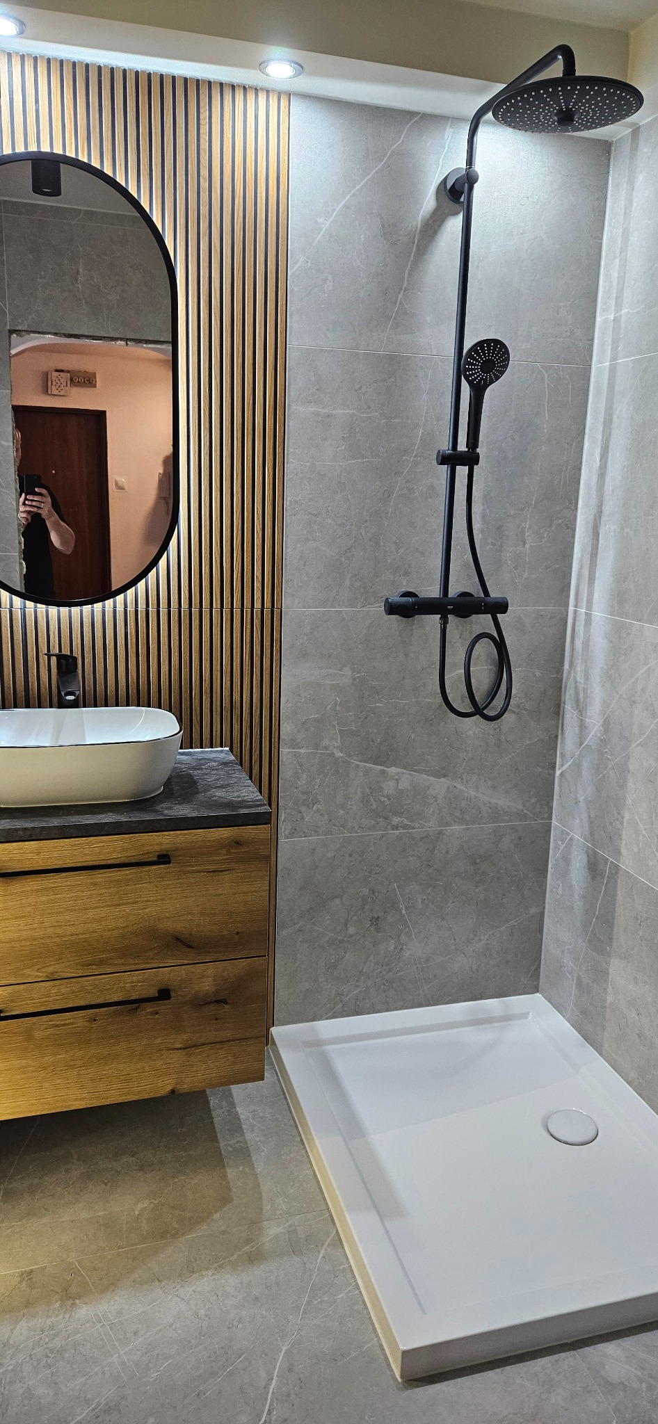Nowoczesna łazienka z drewnianymi lamelami, szarą płytką imitującą marmur i czarną armaturą. Umywalka nablatowa i kabina prysznicowa z brodzikiem.