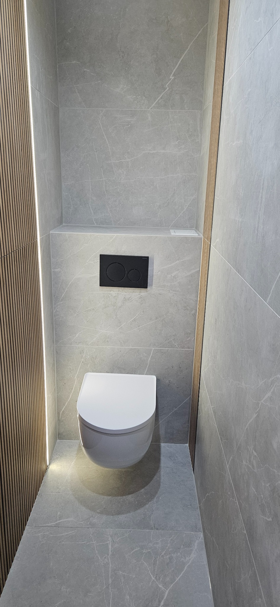Nowoczesna toaleta w szarościach z drewnianymi lamelami i podświetleniem LED. Minimalistyczny design, płytki imitujące marmur, wisząca miska WC i czarny przycisk spłukiwania.