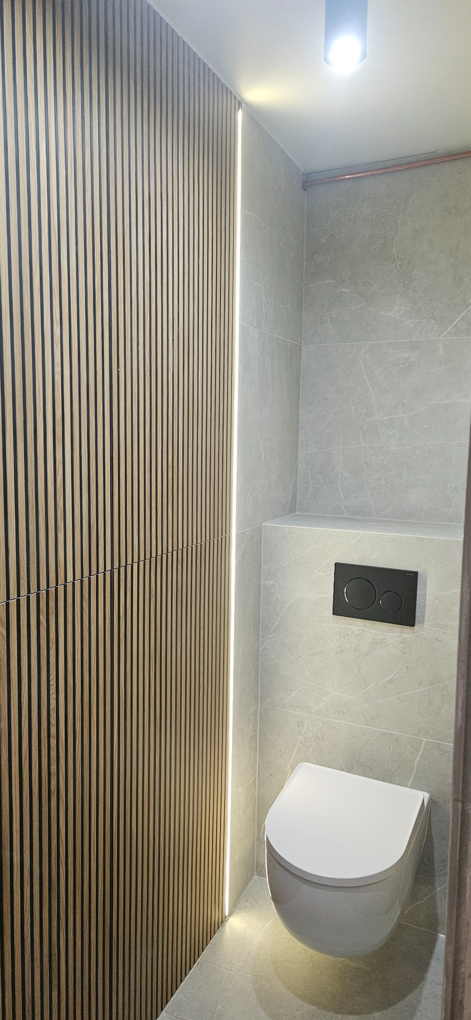 Nowoczesna łazienka z drewnianą ścianą z lameli i podświetleniem LED, szare płytki imitujące marmur, podwieszana toaleta. Minimalistyczny design.