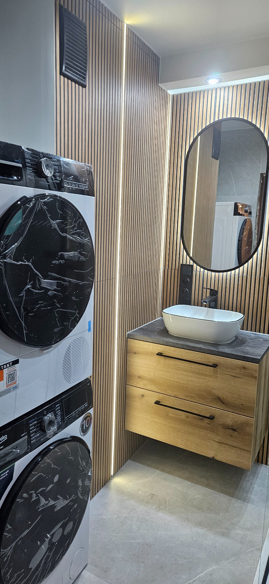 Nowoczesna łazienka z drewnianą ścianą z listew, owalnym lustrem, umywalką nablatową i pralko-suszarką Beko w folii ochronnej. Minimalistyczny design.
