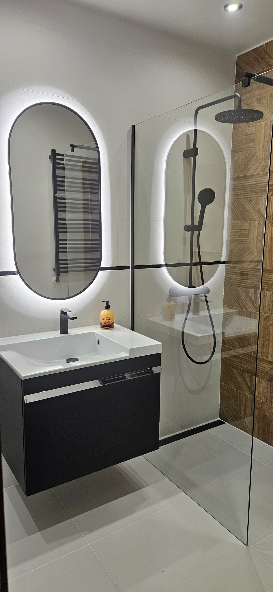 Nowoczesna łazienka z owalnymi lustrami LED, czarną armaturą i kabiną prysznicową ze szklaną ścianką. Minimalistyczny design z drewnopodobnymi płytkami.