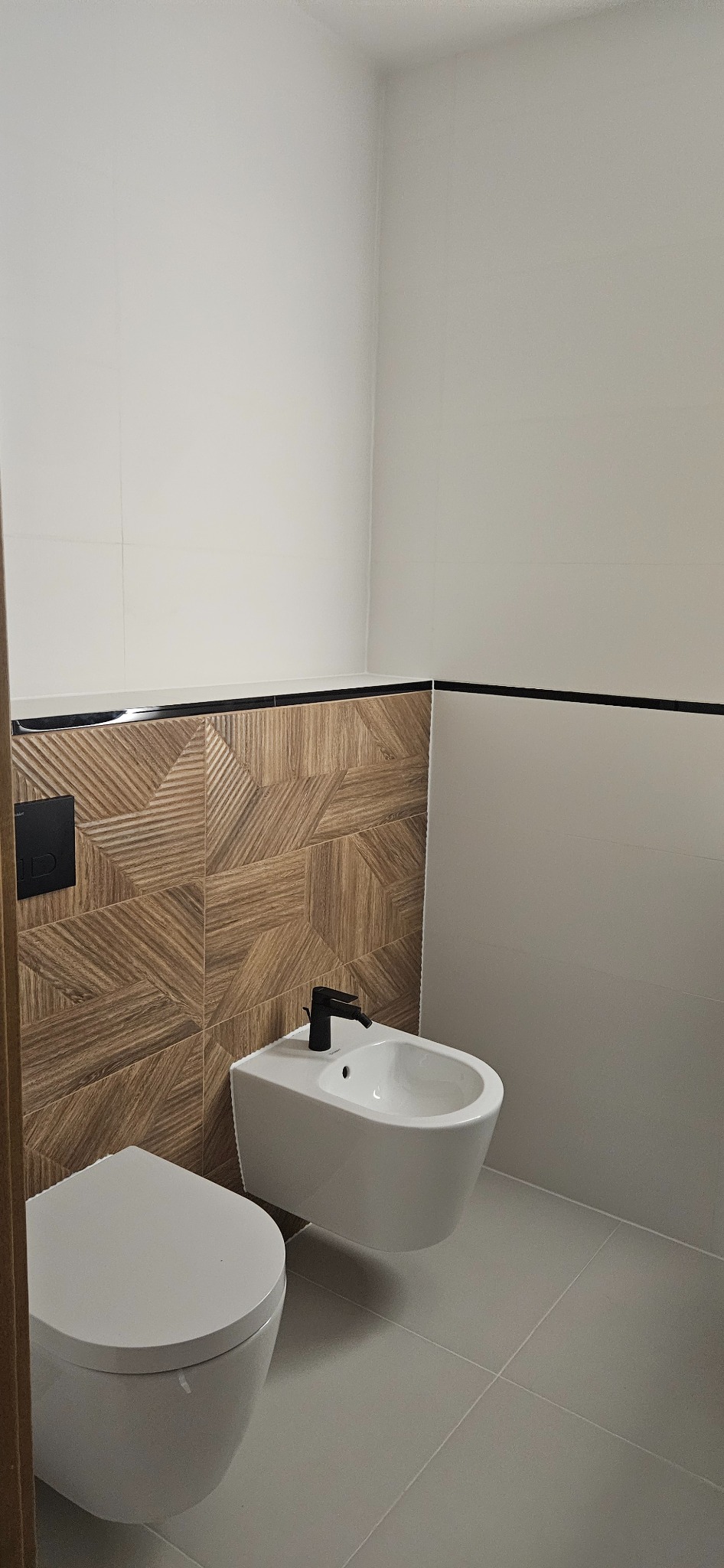 Nowoczesna łazienka z wiszącą toaletą i bidetem. Brązowe płytki imitujące drewno, białe ściany i szara podłoga. Czarna armatura dodaje elegancji.