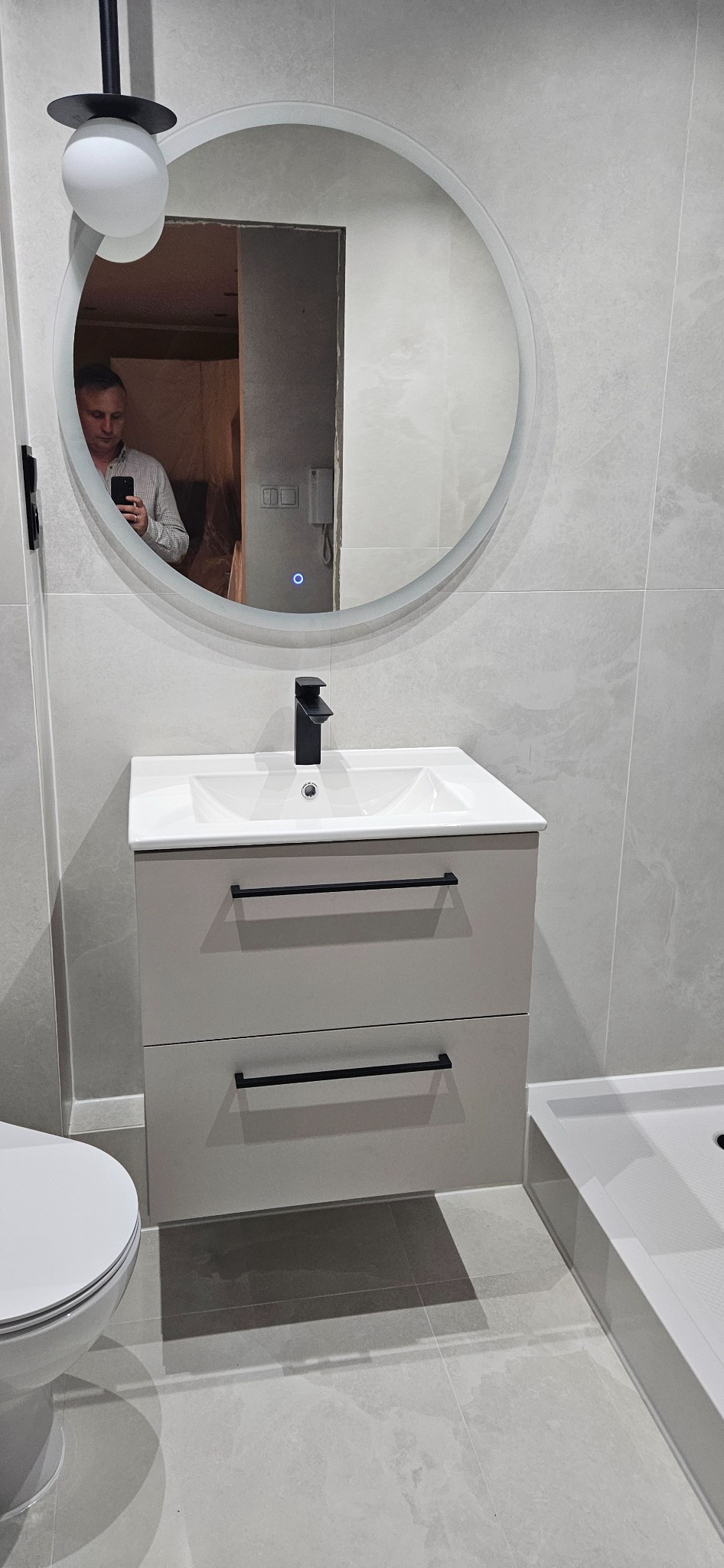 Nowoczesna łazienka w szarościach: umywalka z szafką, okrągłe lustro z podświetleniem i kabina prysznicowa. W lustrze odbija się osoba robiąca zdjęcie.