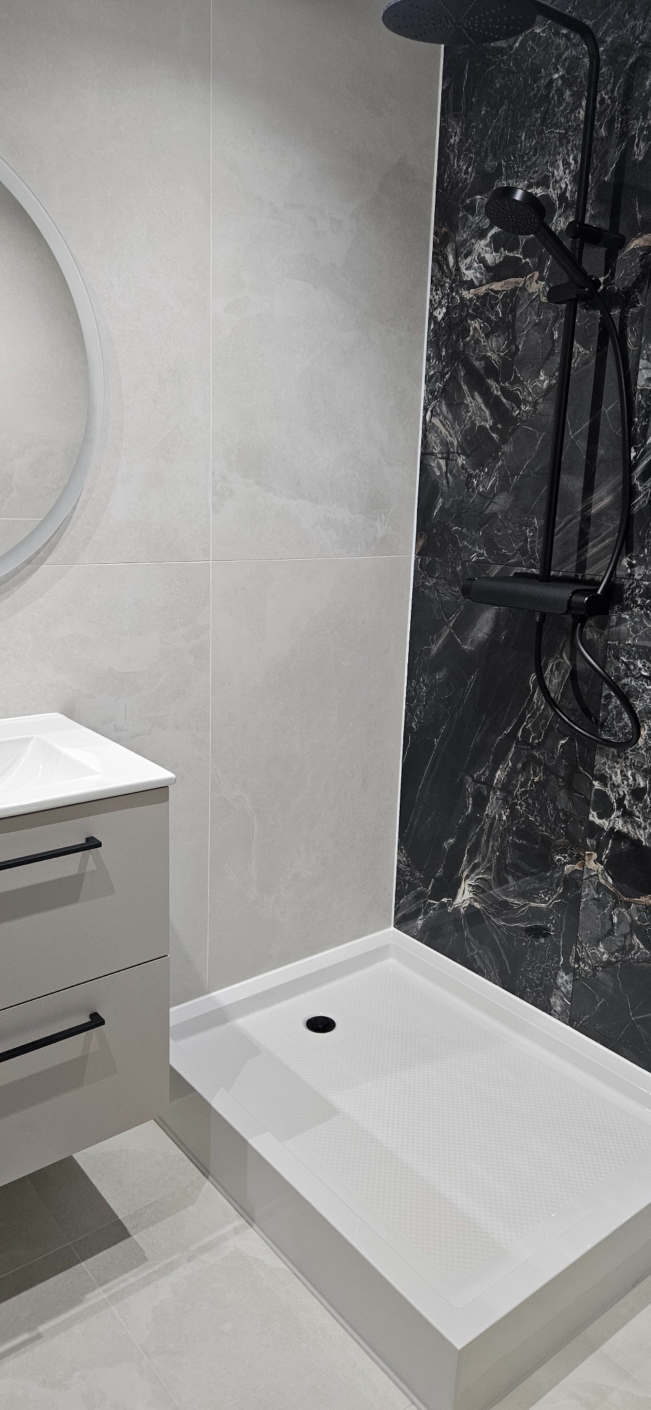 Nowoczesna łazienka z kabiną prysznicową typu walk-in, umywalką z szafką w odcieniach szarości i czarną armaturą na tle marmurowej ściany.