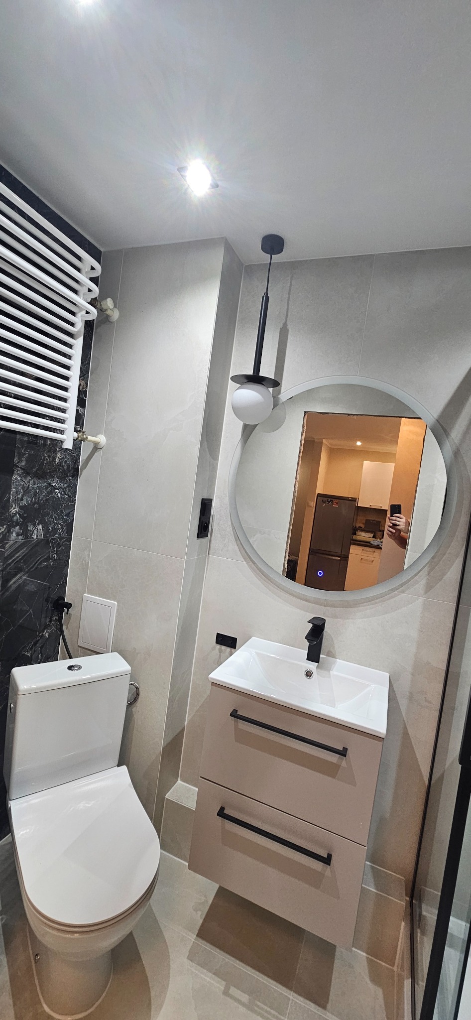 Nowoczesna łazienka z beżowymi płytkami, wiszącą szafką z umywalką i okrągłym lustrem, w którym odbija się fragment kuchni. Minimalistyczny design, czarne akcenty.