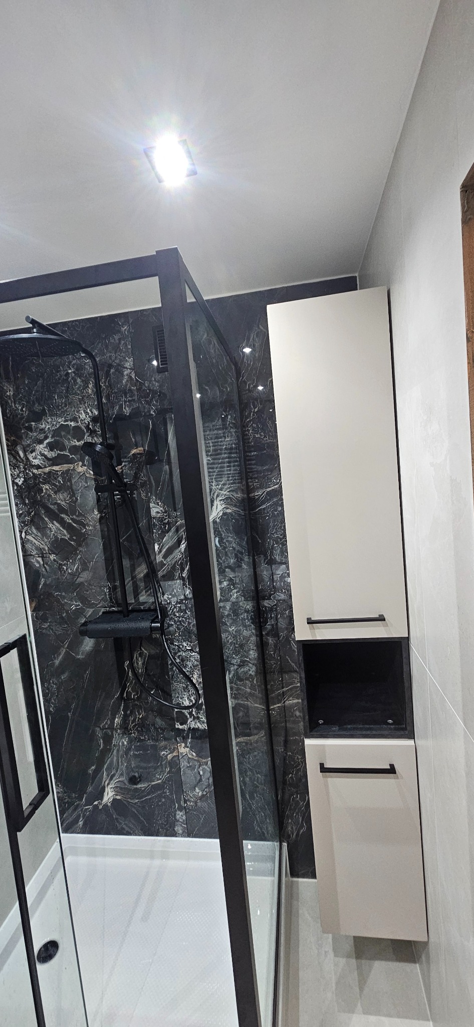 Nowoczesna kabina prysznicowa z czarną armaturą i marmurową ścianą, uzupełniona o białą szafkę łazienkową z czarnymi uchwytami. Minimalistyczny design.