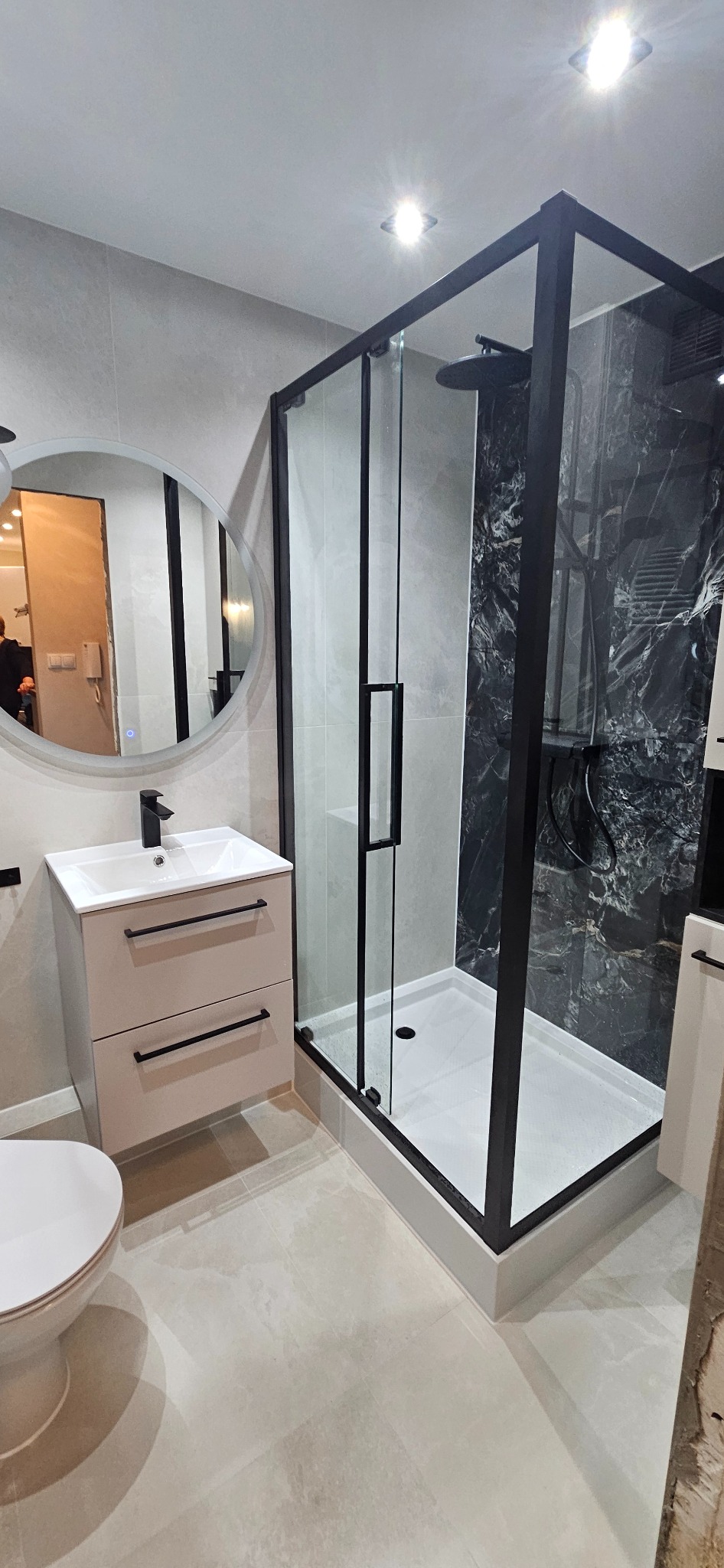 Nowoczesna łazienka z kabiną typu walk-in z czarnymi profilami, umywalką z szafką w beżowym odcieniu i okrągłym lustrem z podświetleniem LED.
