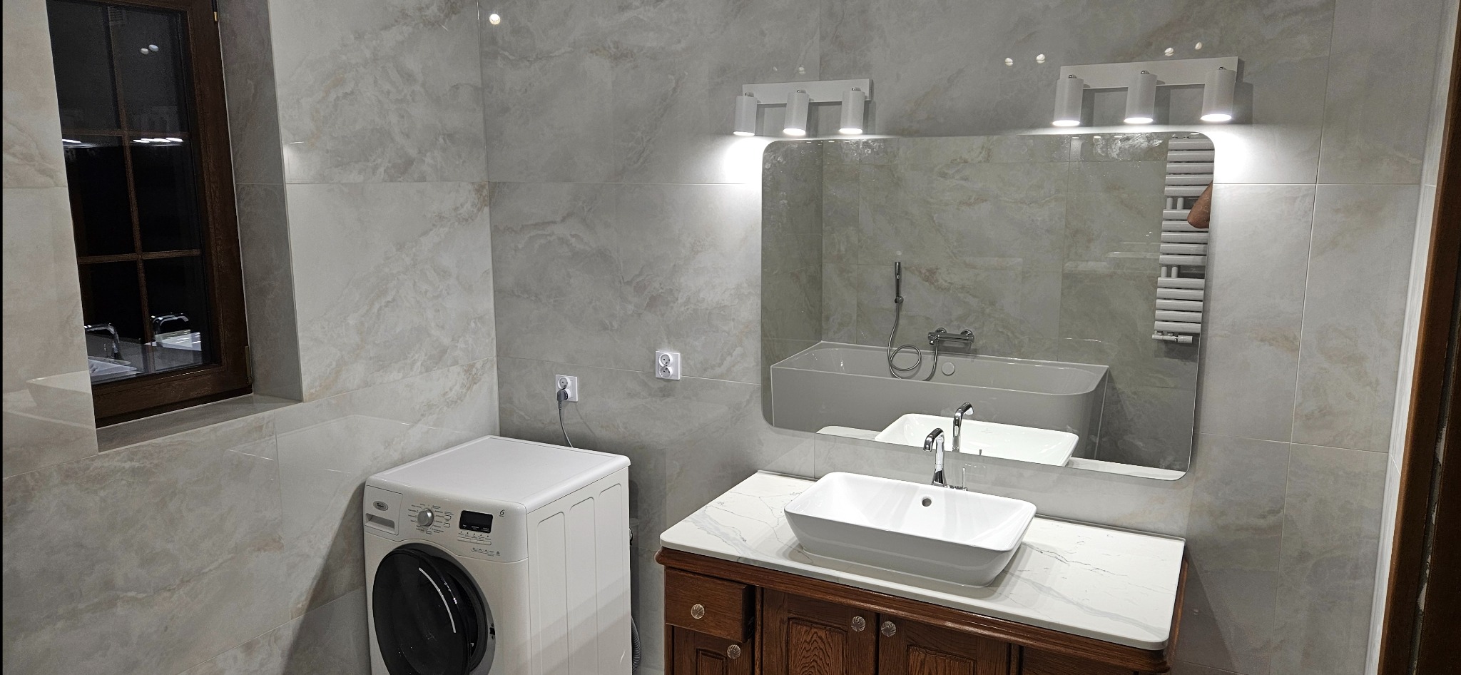 Wykończona łazienka w beżowych płytkach z pralką, umywalką na szafce z marmurowym blatem i lustrem z oświetleniem. Widoczne gniazdka elektryczne i okno.