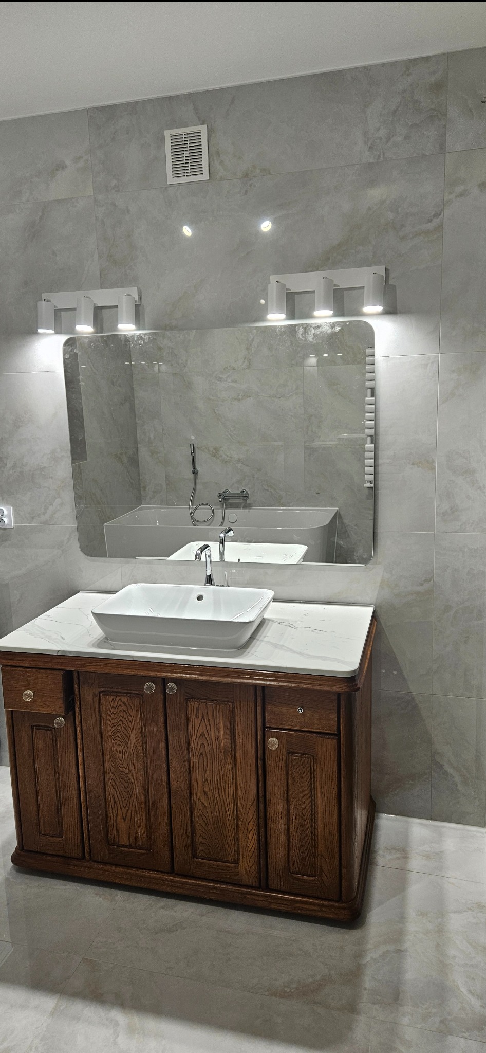 Wykończona łazienka z drewnianą szafką pod umywalką, marmurowym blatem i dużym lustrem. Widoczne oświetlenie punktowe i płytki w odcieniach szarości.