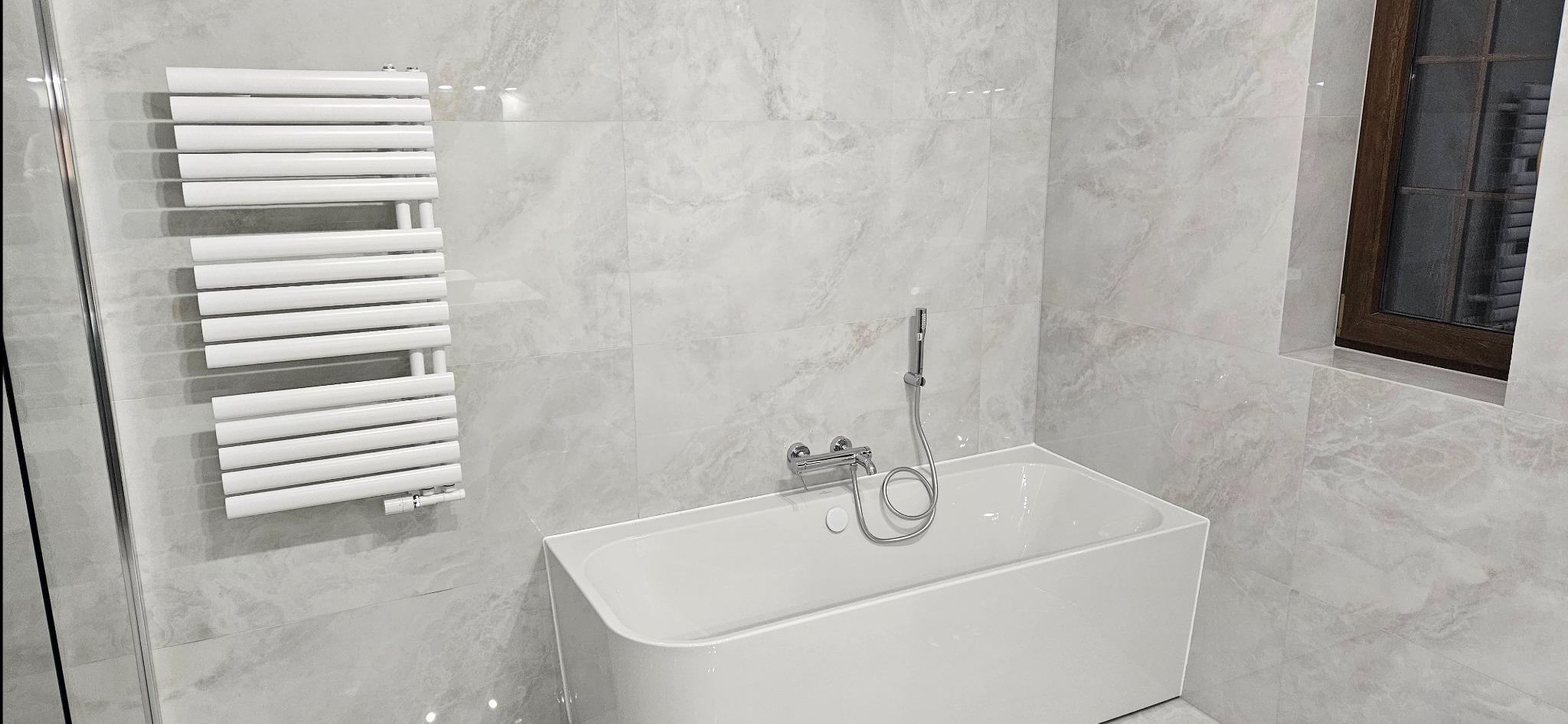 Biała wanna z baterią i słuchawką prysznicową na tle marmurowych płytek. Obok grzejnik drabinkowy i okno z brązową ramą. Minimalistyczna aranżacja łazienki.