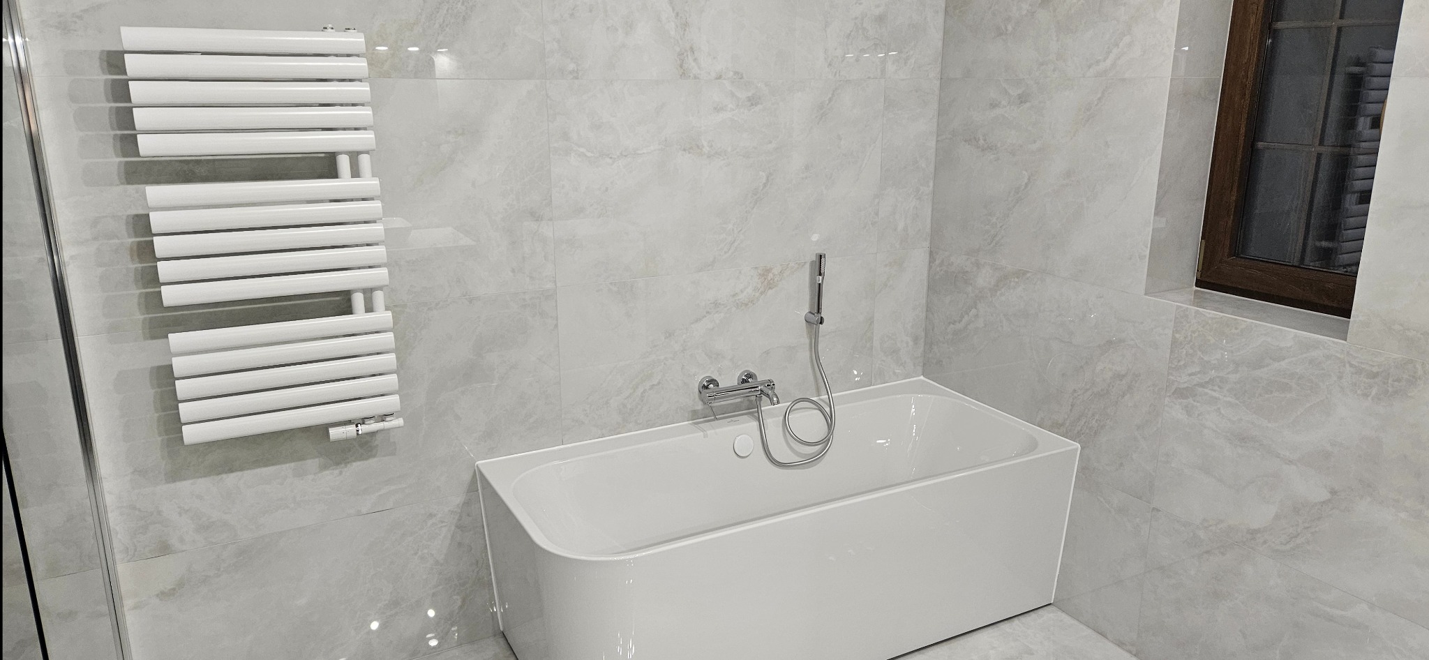 Nowoczesna łazienka z wanną, grzejnikiem drabinkowym i oknem. Ściany i podłoga wyłożone płytkami imitującymi marmur w odcieniach szarości. Minimalistyczny design.