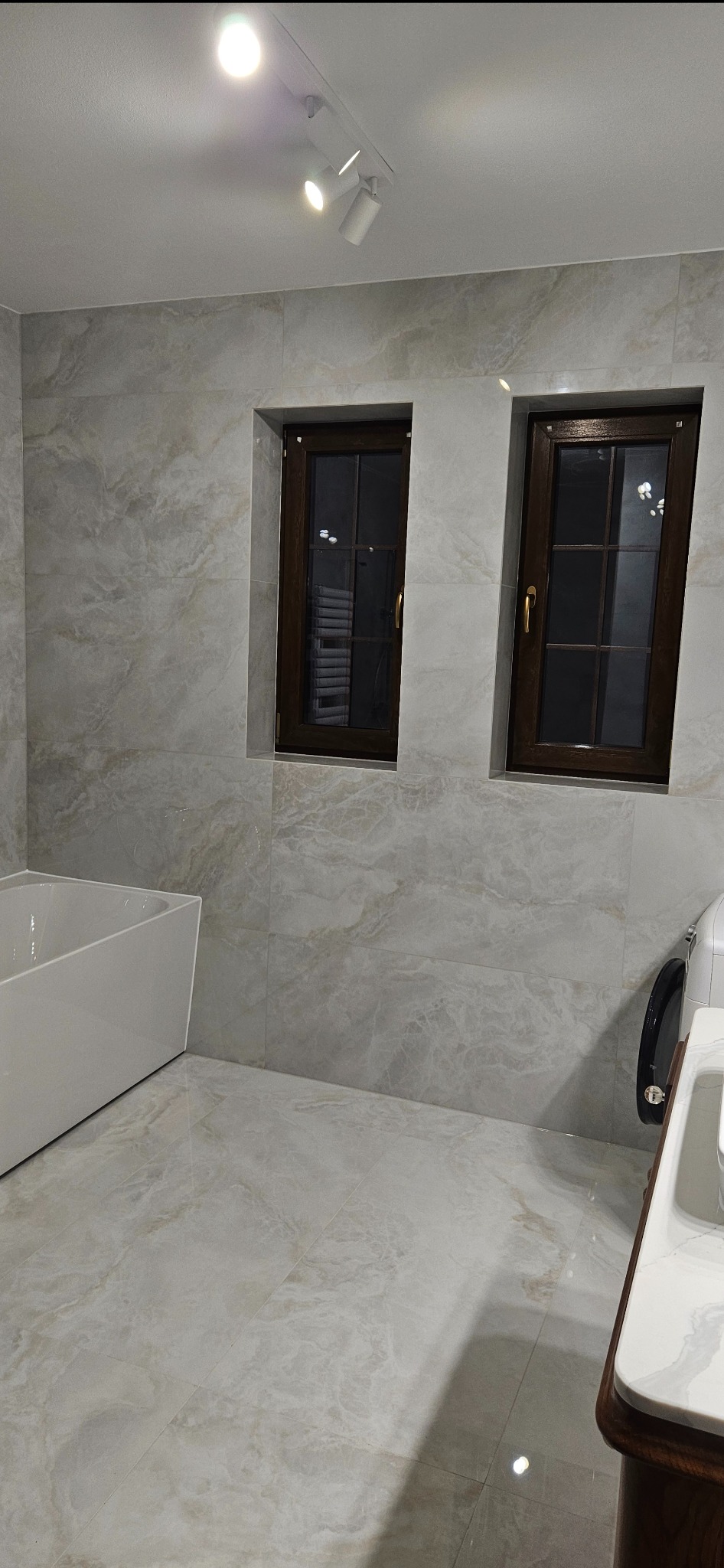 Wykończona łazienka z marmurowymi płytkami, brązowymi oknami i nowoczesną wanną. Widoczne oświetlenie sufitowe i fragment umywalki z szafką.