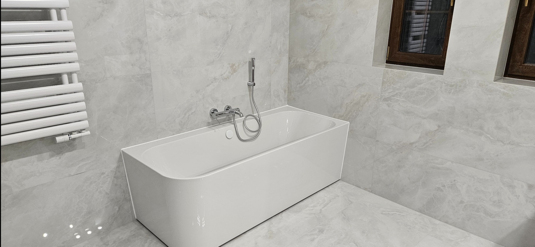 Nowoczesna łazienka z wanną, grzejnikiem drabinkowym i marmurowymi płytkami. Minimalistyczny design, chromowana armatura, okno z ciemną ramą.