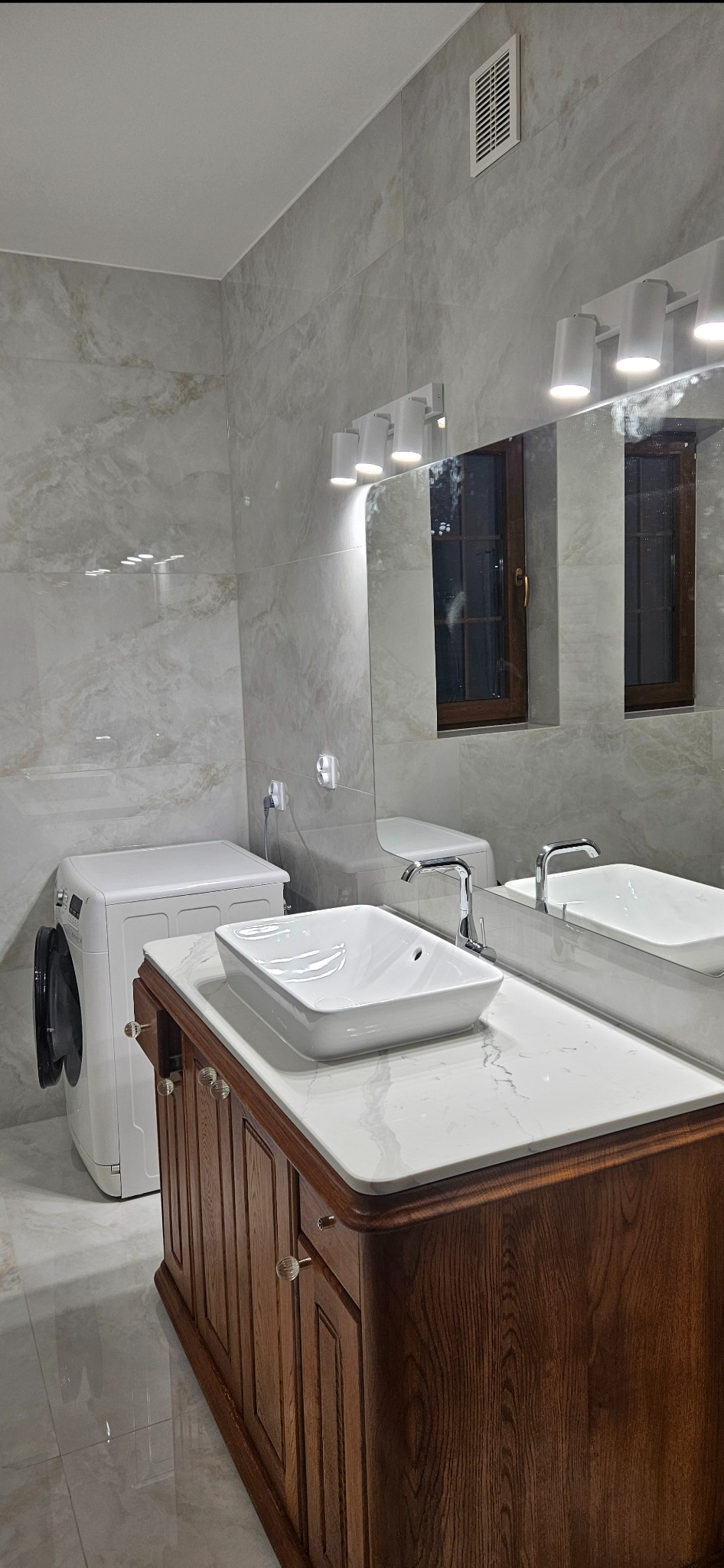 Nowoczesna łazienka z marmurowymi płytkami, drewnianą szafką pod umywalką i pralką. Lustro z oświetleniem punktowym. Minimalistyczny design.