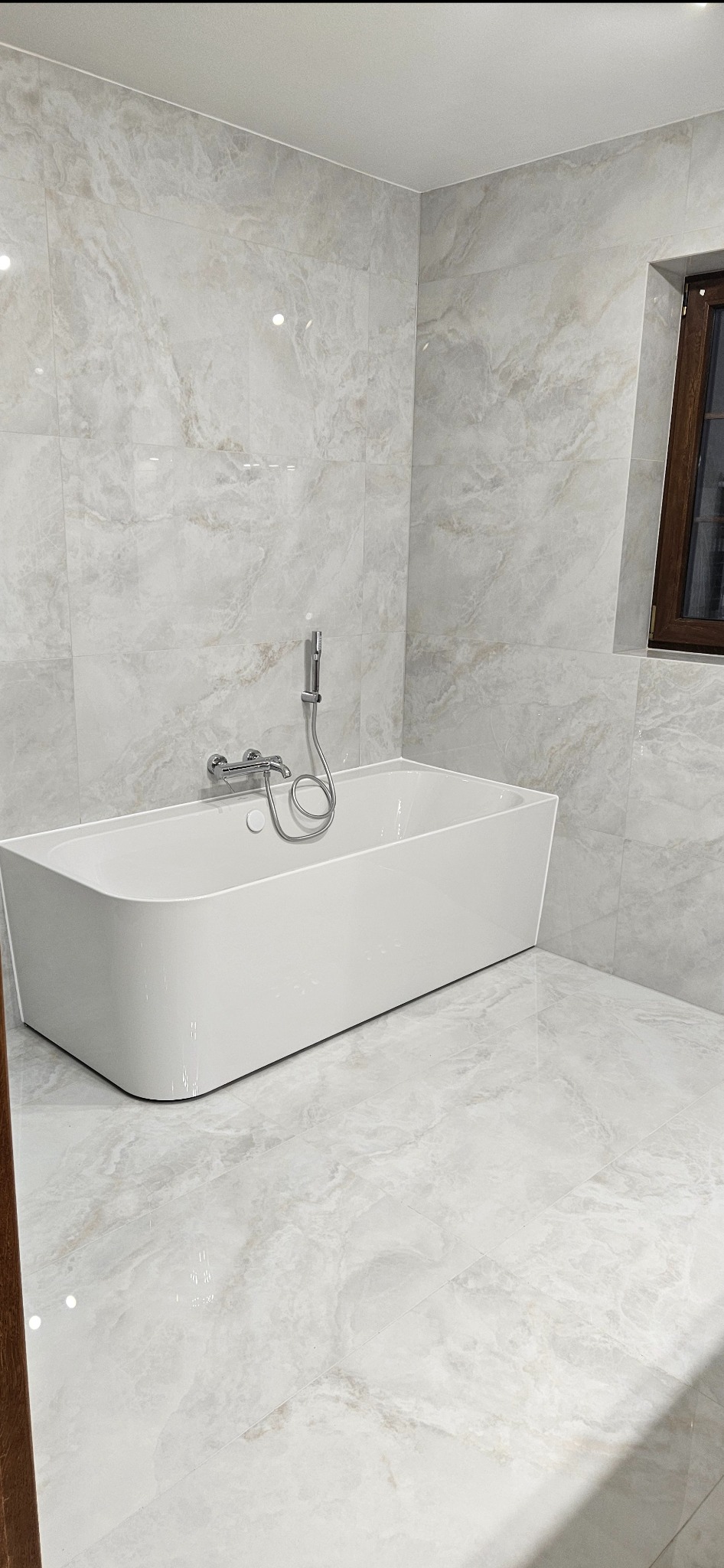 Nowoczesna, jasna łazienka z wolnostojącą wanną i marmurowymi płytkami. Bateria wannowa chromowana, okno z ciemną ramą. Minimalistyczny design.