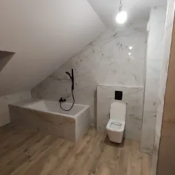 Ukończona łazienka na poddaszu z wanną obudowaną płytkami imitującymi marmur, czarną armaturą i podwieszaną toaletą, wykończona panelami podłogowymi w odcieniach drewna.