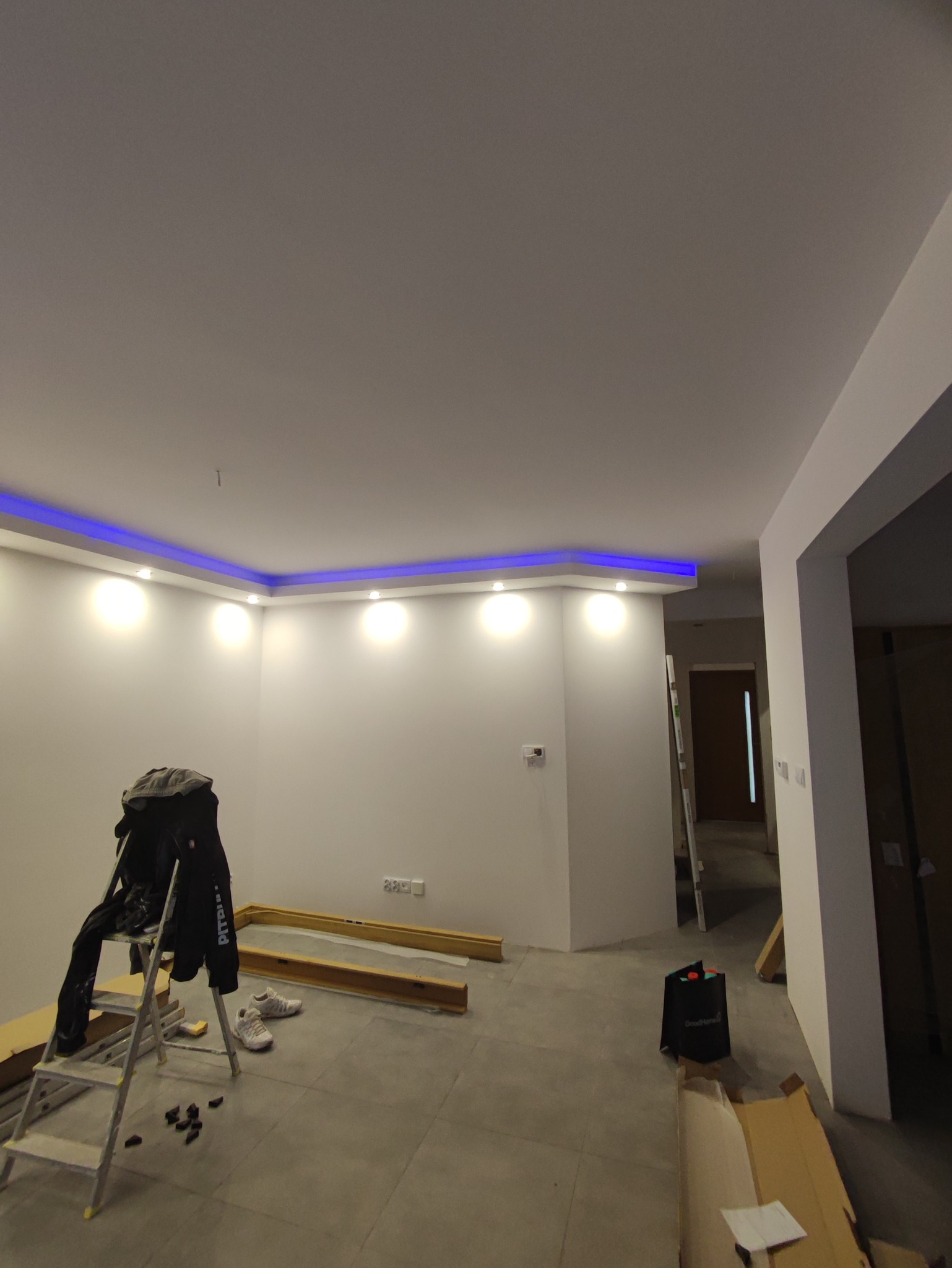 Pokój w trakcie prac wykończeniowych: gładkie, białe ściany z zamontowanym oświetleniem LED na suficie, drabina malarska z kurtką, panele podłogowe i kartony na podłodze.