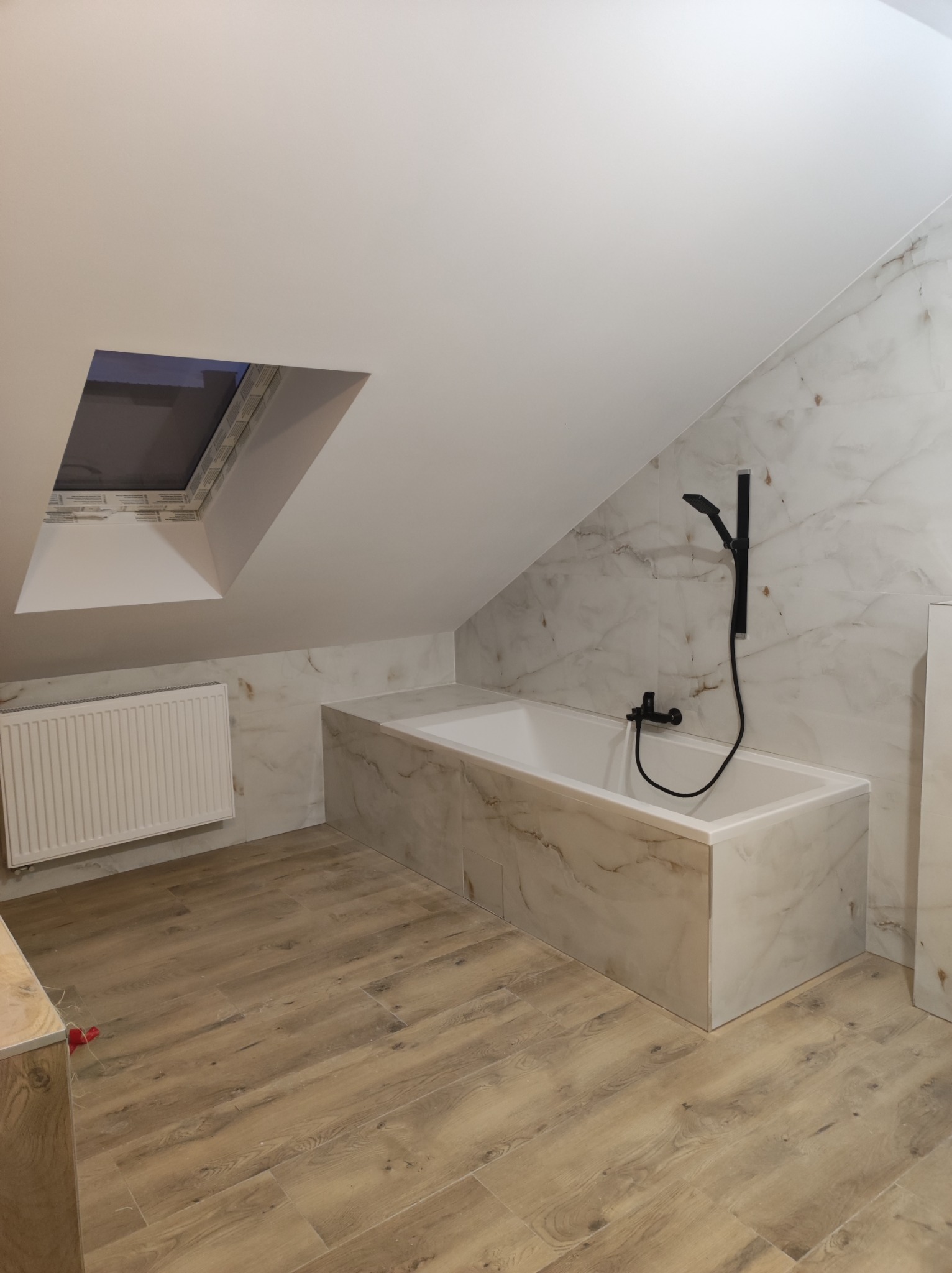 Wykończona łazienka na poddaszu ze skośnym sufitem, białą wanną obudowaną płytkami imitującymi marmur, czarną armaturą i drewnopodobną podłogą. Widoczne okno dachowe i grzejnik.