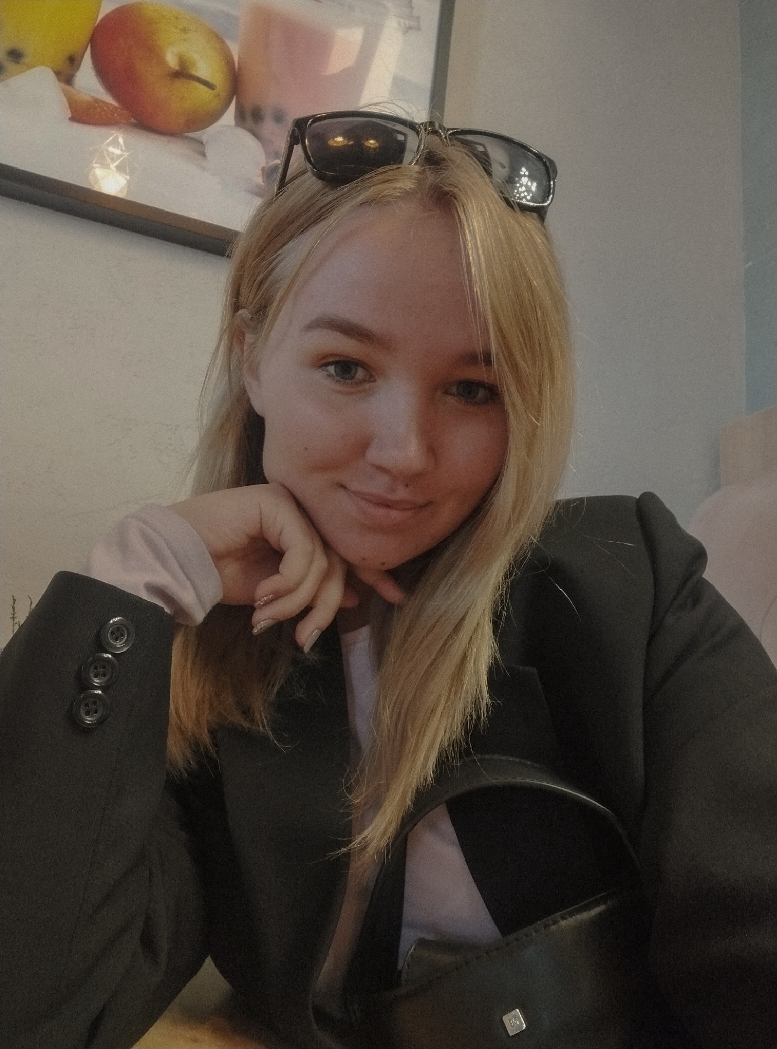 Portret kobiety w czarnej marynarce, z okularami na głowie, uśmiechniętej, z dłonią podpartą pod brodą, w tle widoczny obrazek z owocami i napojami.