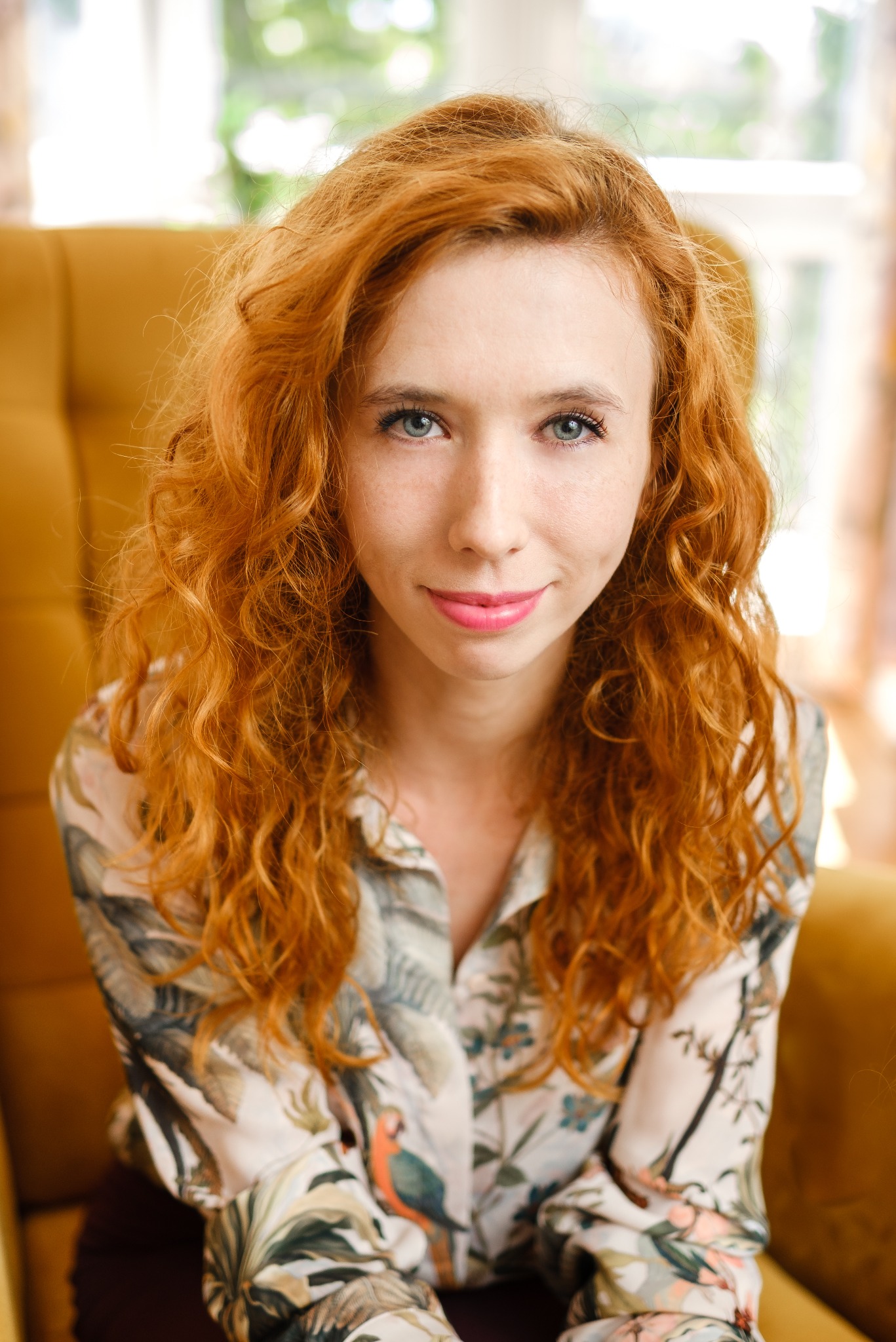 Portret kobiety z rudymi kręconymi włosami i niebieskimi oczami, siedzącej w fotelu w bluzce w tropikalne wzory, z delikatnym uśmiechem.