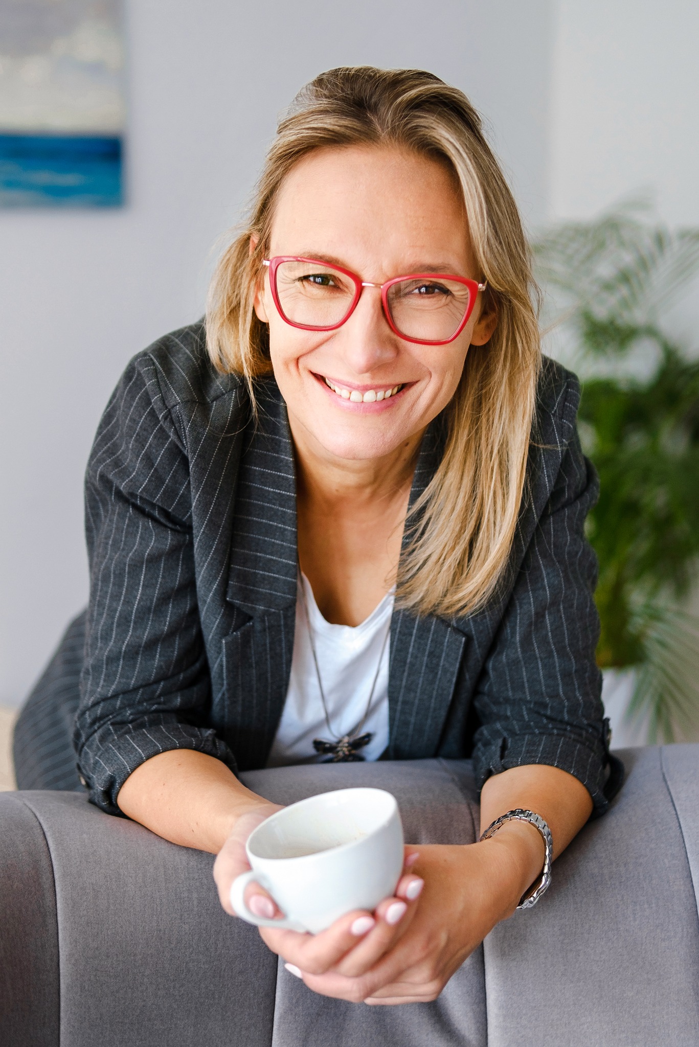 Uśmiechnięta kobieta w szarym żakiecie w paski i czerwonych okularach trzymająca biały kubek z napojem, opierająca się o szary fotel, w tle roślina doniczkowa i obraz z motywem morskim.